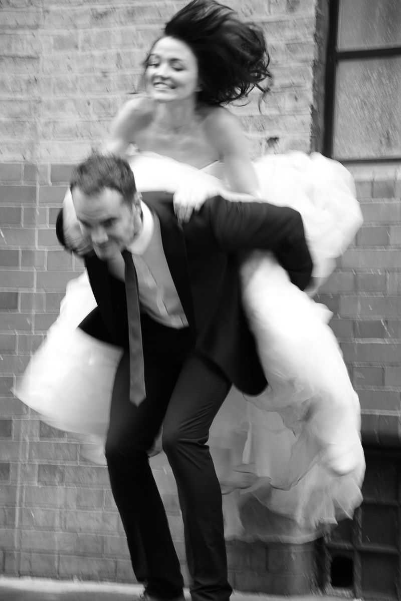 Black and white photo of a groom carrying his laughing bride on his back in a wedding dress outdoors.