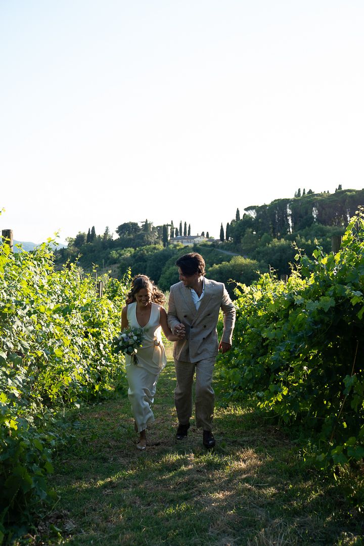 Bride and groom walk hand in hand through lush vineyard rows on their wedding day in Umbria, Italy. At Casale San Cristoforo venue.