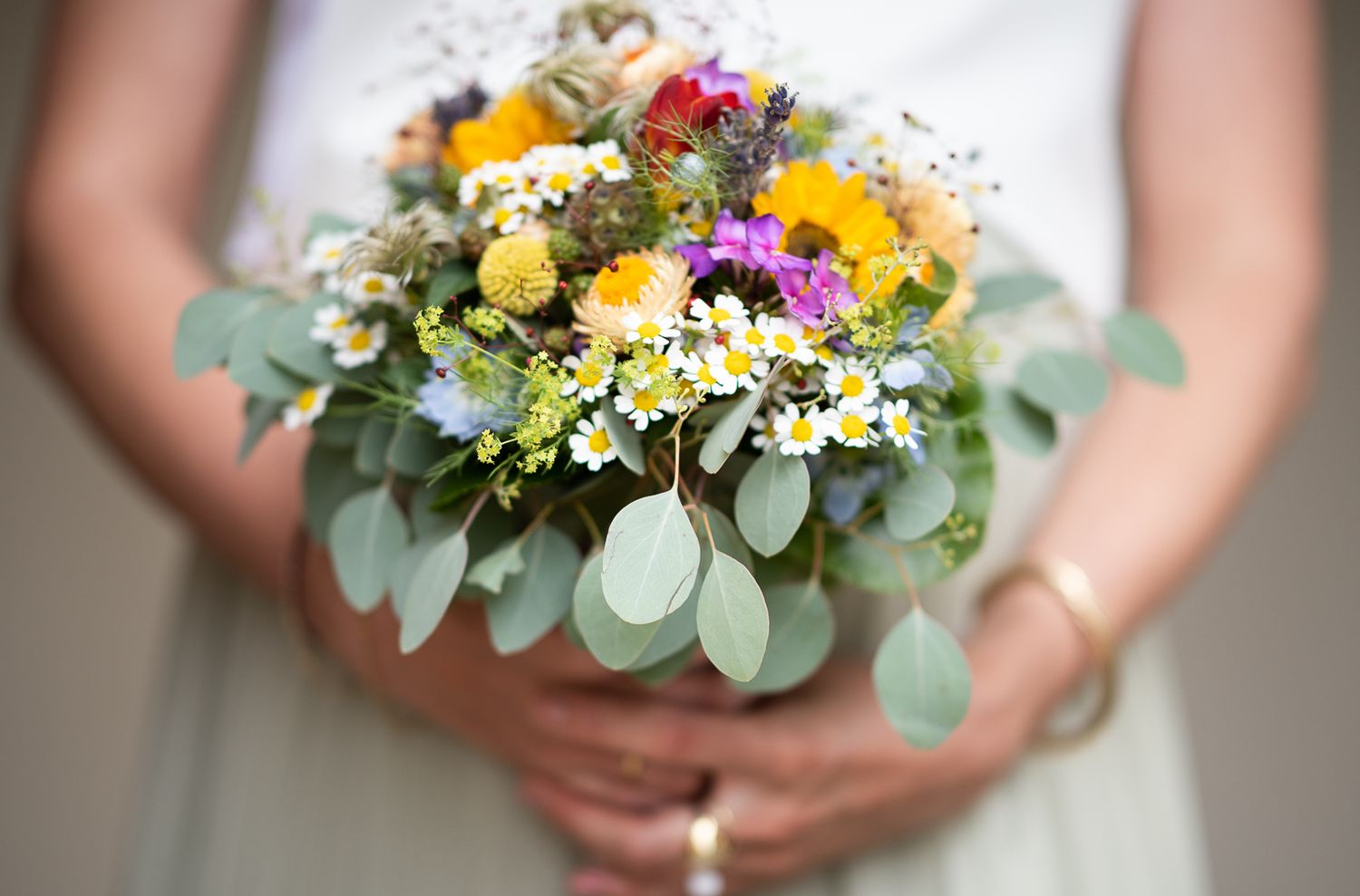 Close-up view of hands holding a vibrant wedding bouquet with yellow, pink and white wildflowers and eucalyptus leaves.