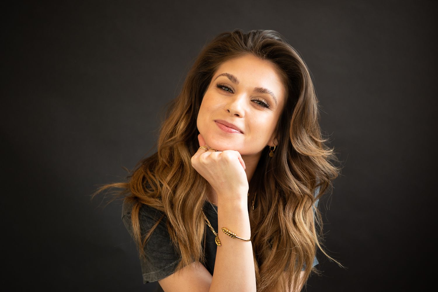 A woman with flowing brown hair poses against a dark background in a glamorous photoshoot.