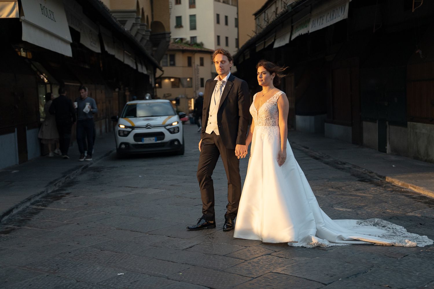 A bride and groom walk along a cobblestone street at sunset while a photographer captures their special moment.