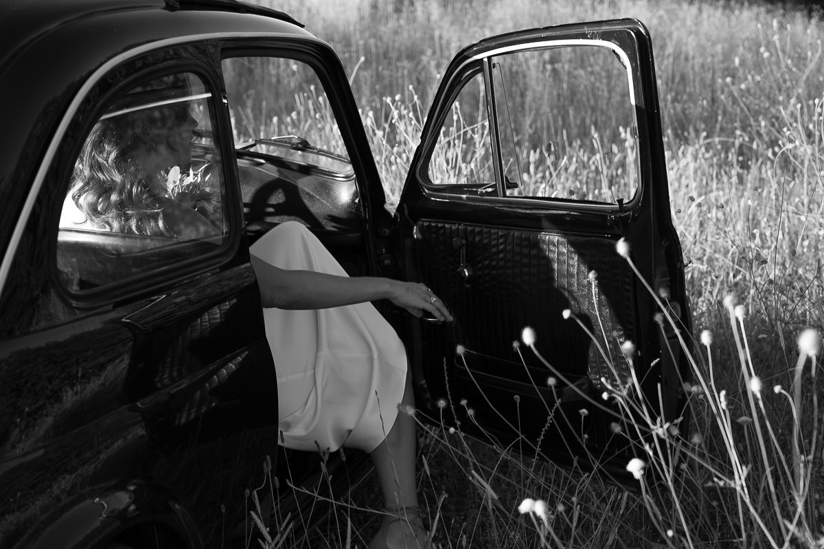 Black and white photo of a woman in a dress leaning into a vintage car door in a wild meadow.