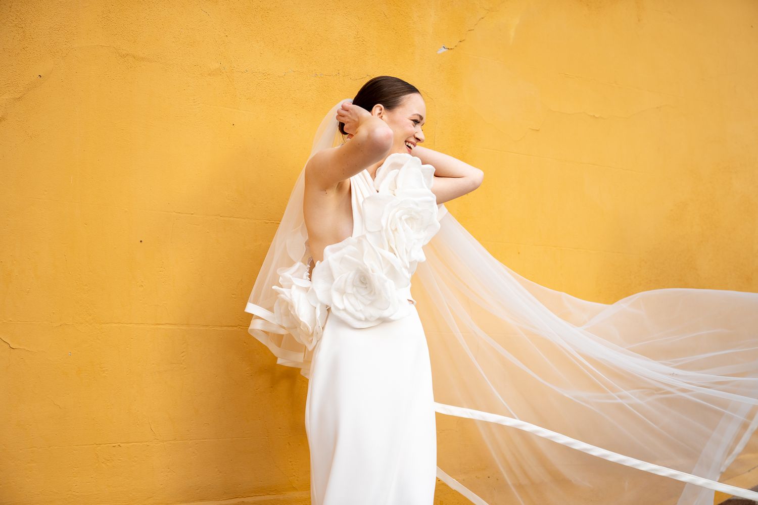 A bride in a flowing white wedding dress and veil stands against a vibrant yellow wall, creating an elegant contrast.