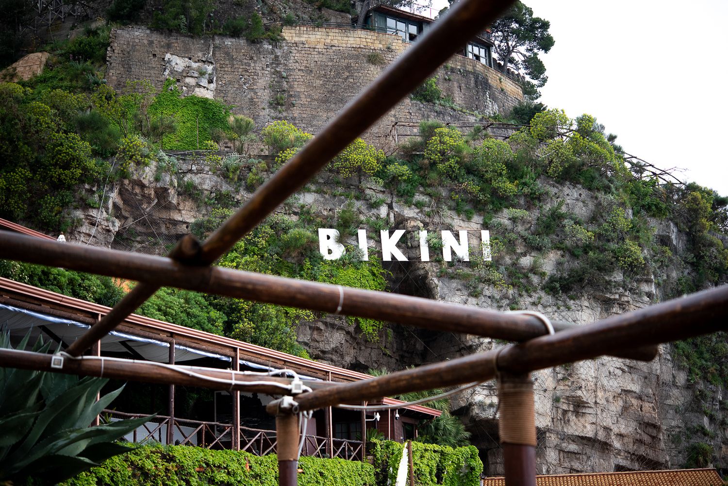 View of BIKINI text sign mounted on rocky cliff wall with rustic beams in foreground.