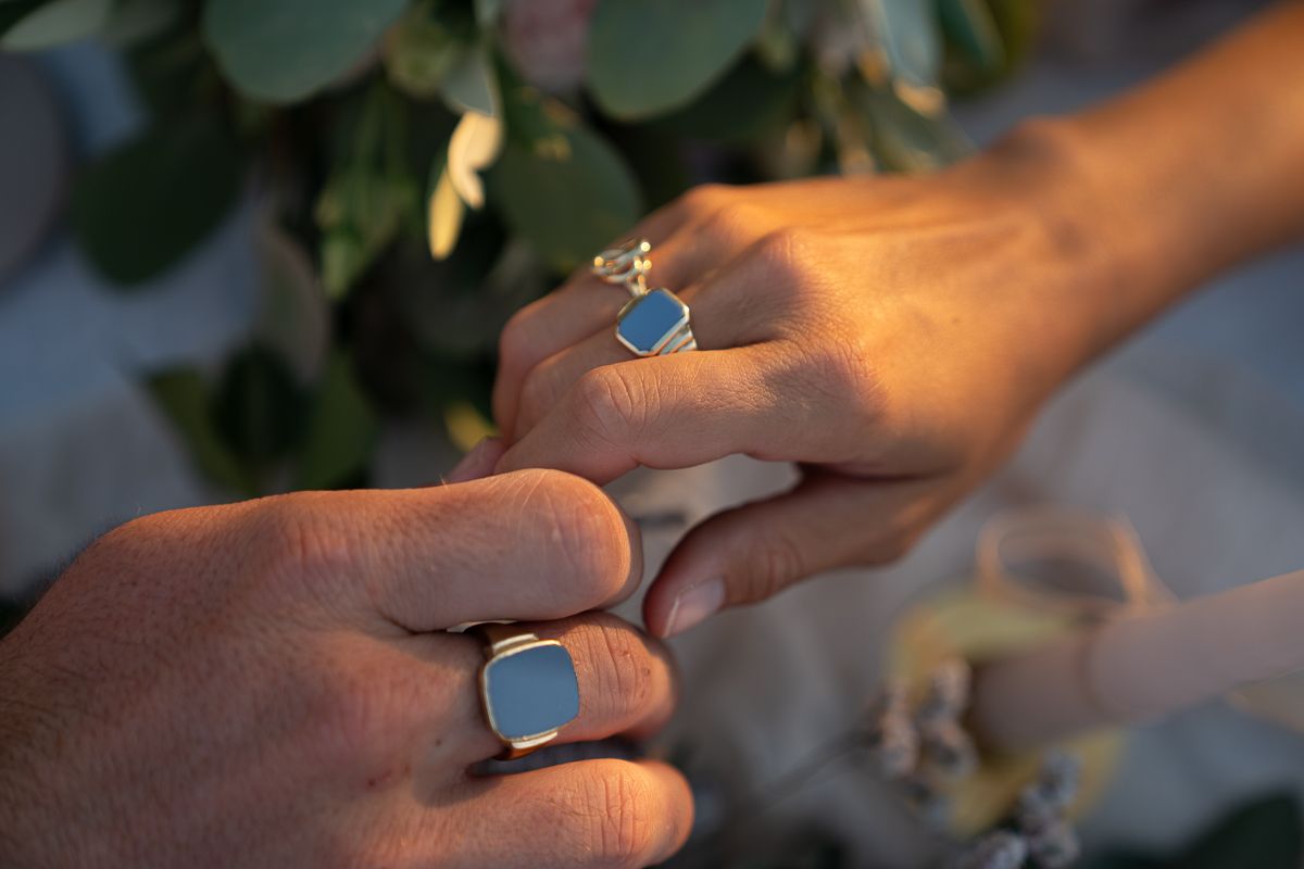 Two hands adorned with matching blue stone gold rings touch gently against a floral background on elopement day.