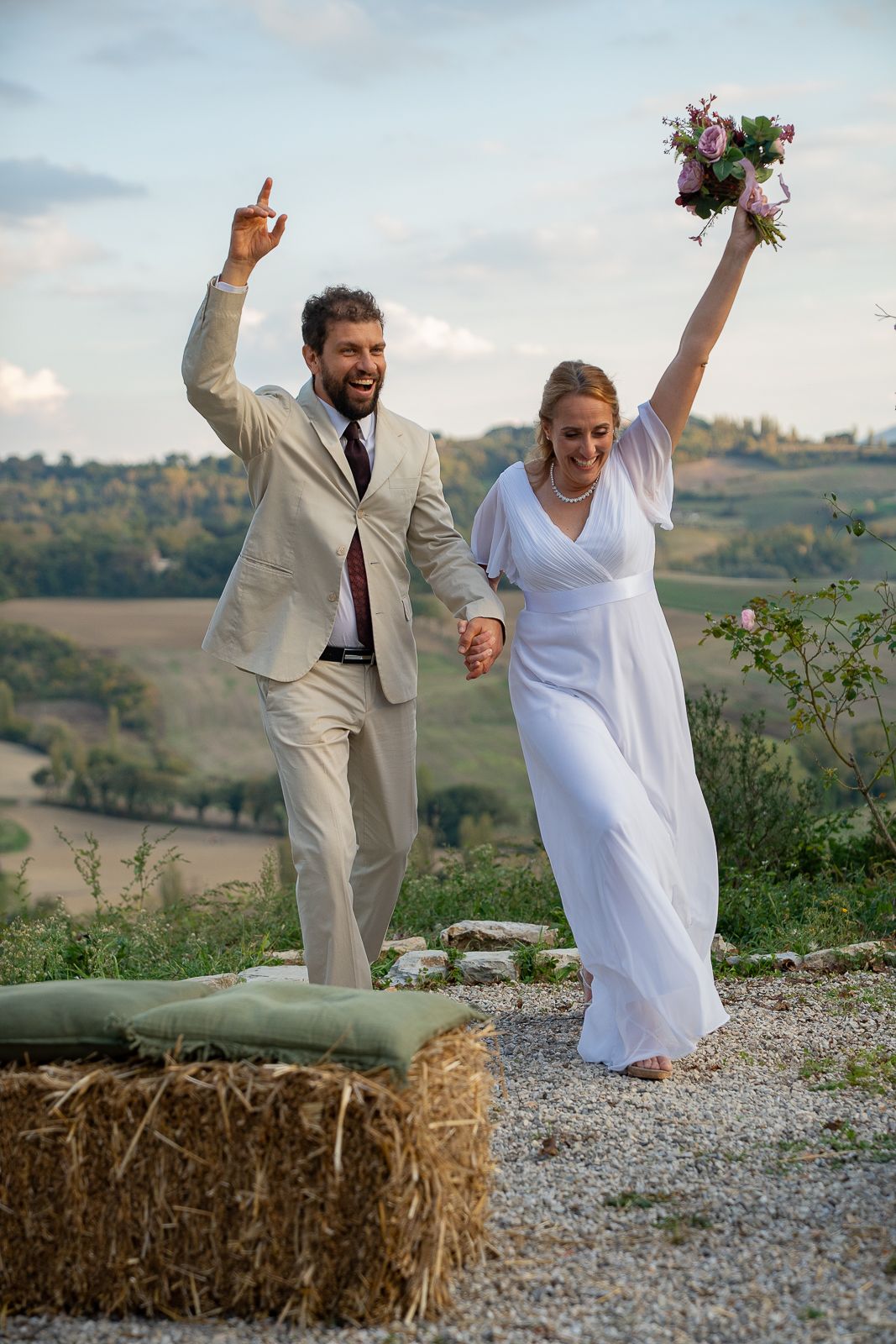A couple in wedding attire celebrates joyfully on a hilltop with hay bales and countryside views in the background at Casale San Cristoforo