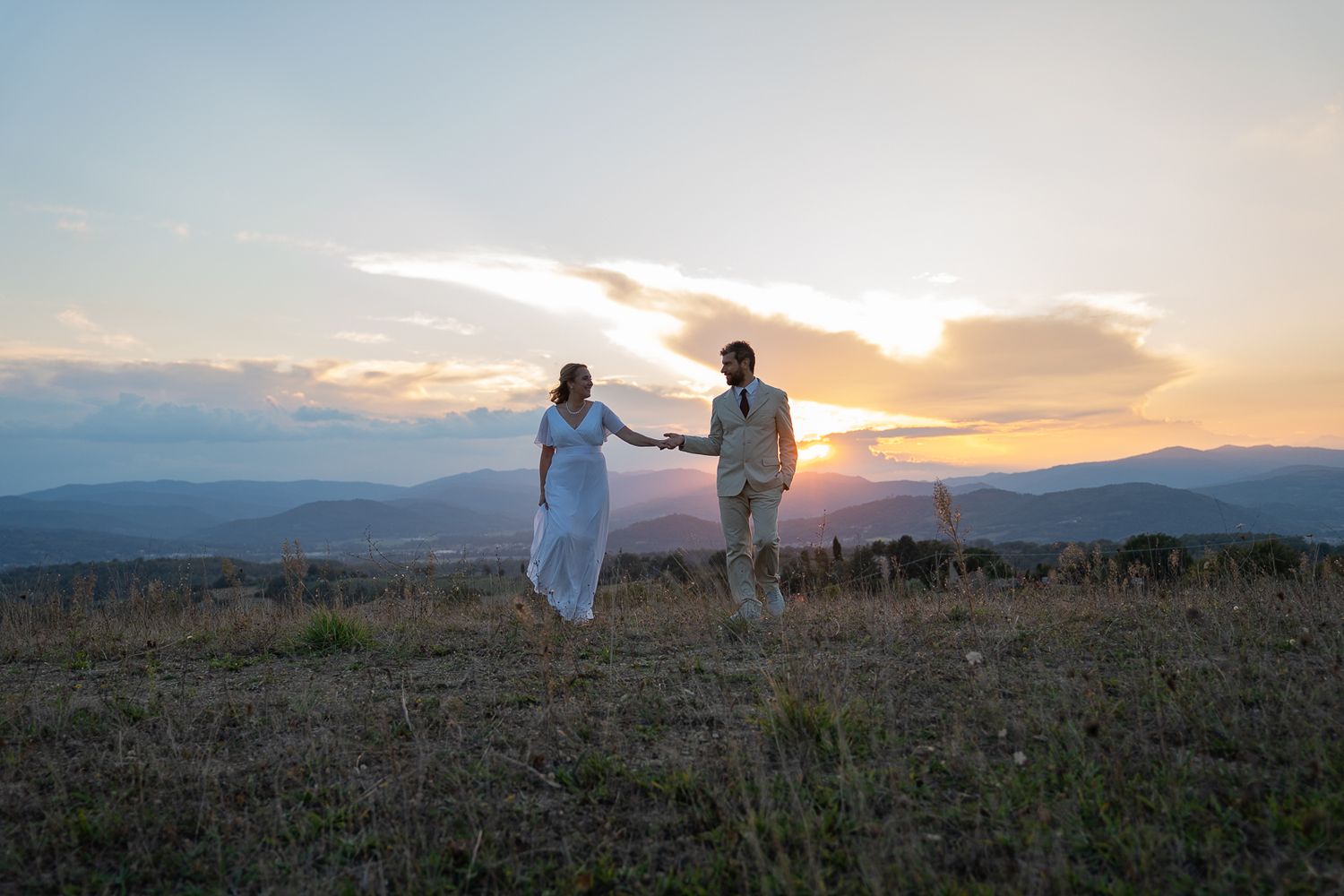 A couple holds hands on a hilltop during a golden sunset over mountain ranges in the distance.