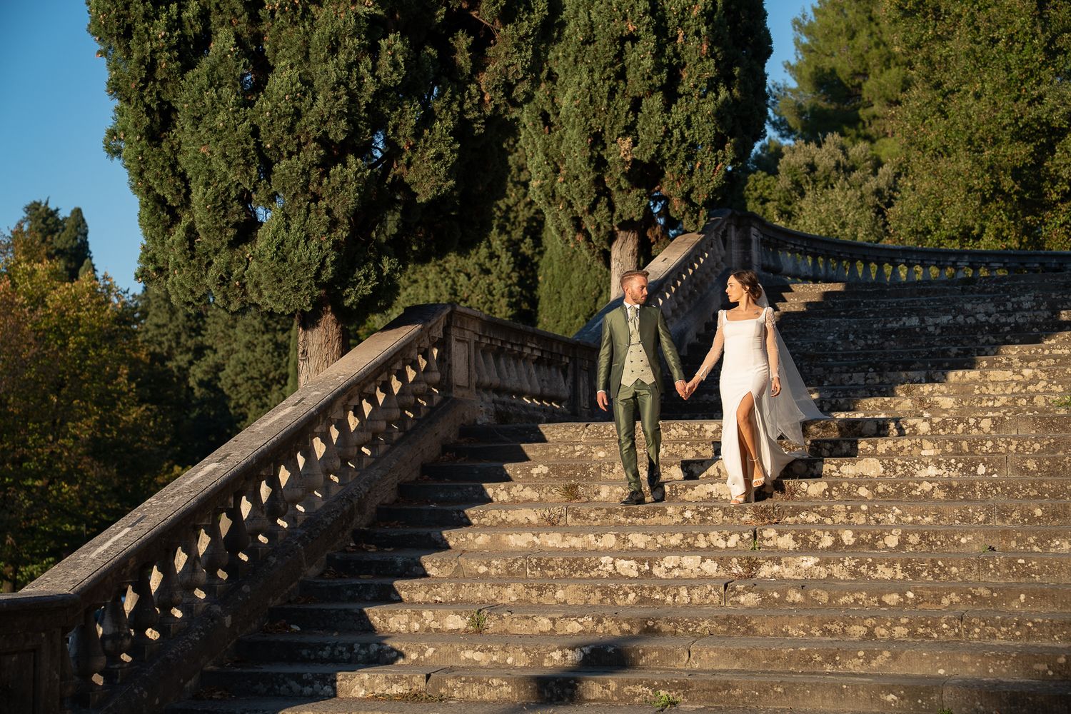 A figure descends stone steps in a garden surrounded by tall cypress trees in warm afternoon sunlight.