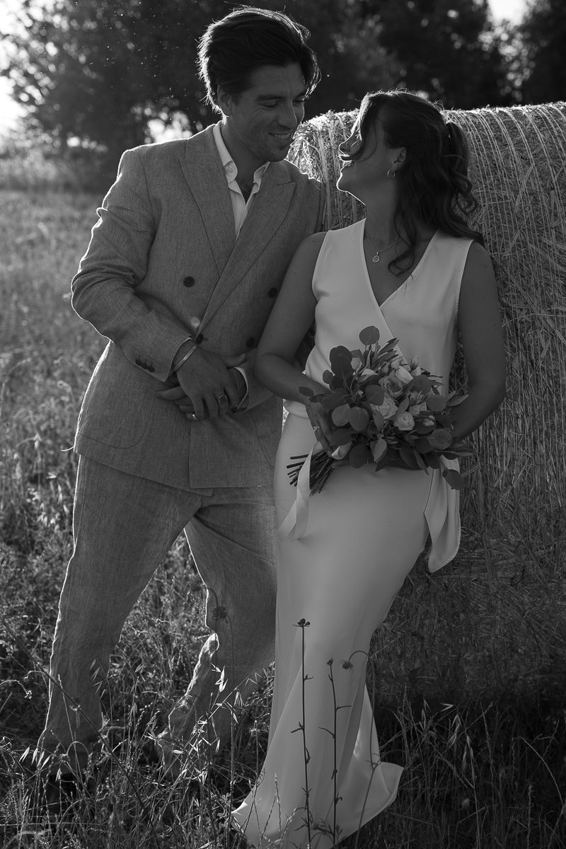Black and white photo of a bride and groom leaning against a hay bale in a field, smiling at each other shot by Friderika Wollweber.