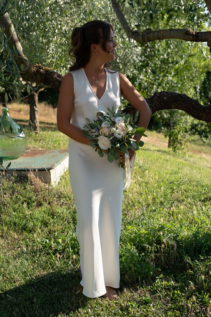 Bride in white sleeveless gown holding floral bouquet stands barefoot in lush green garden under olive trees at Casale San Cristoforo Italy