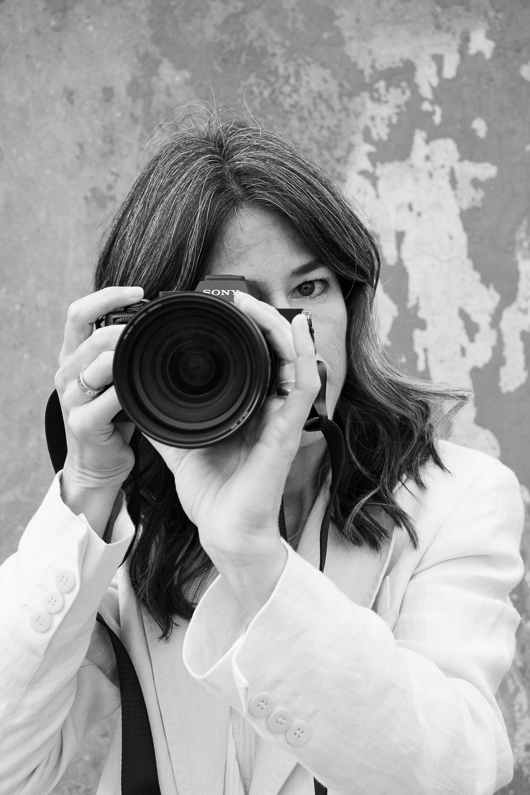 Black and white photo of a woman holding a Sony camera up to her face, wearing a white blazer.