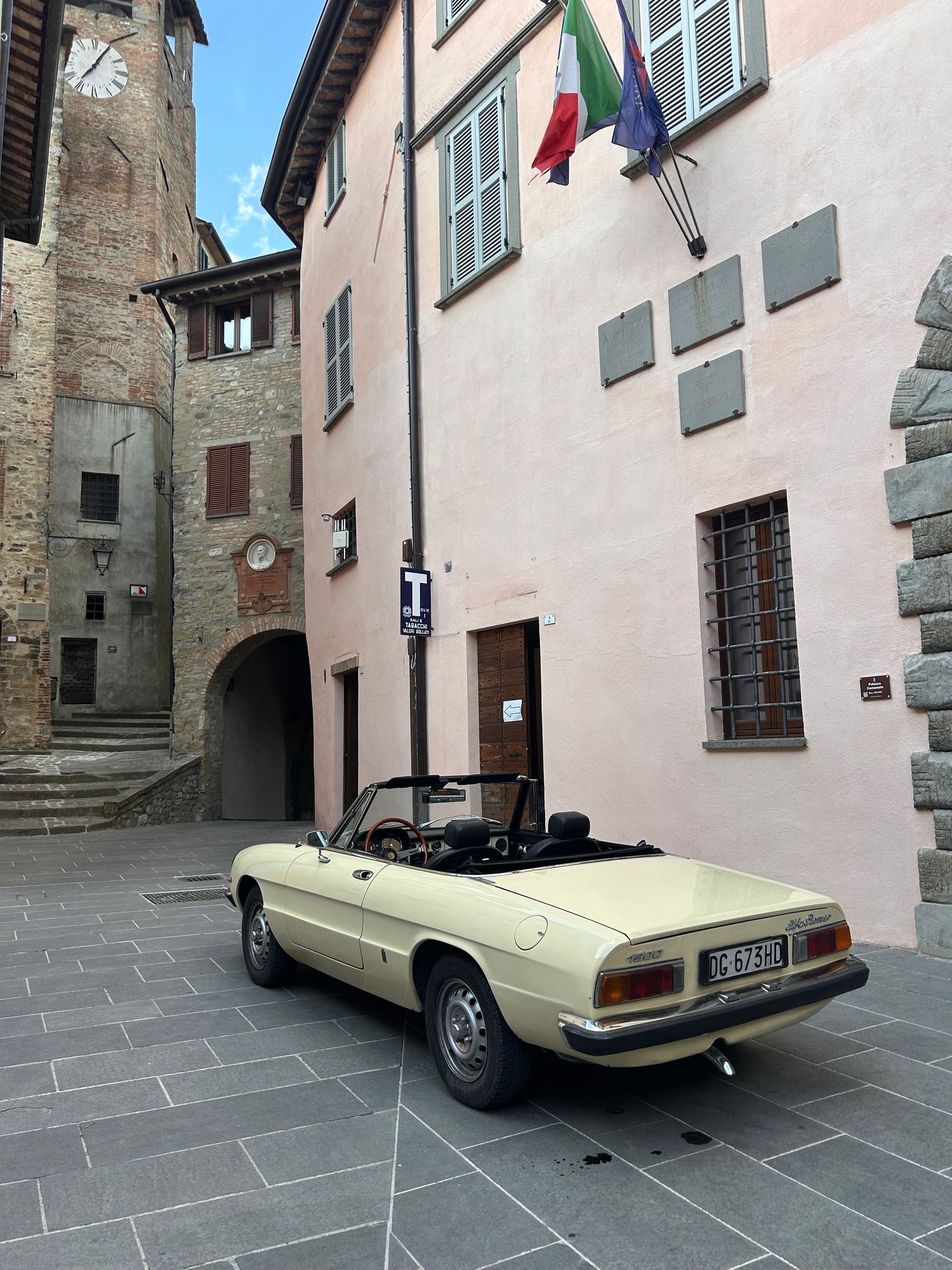 Vintage cream convertible car parked in a historic Italian piazza with stone buildings and Italian flag.