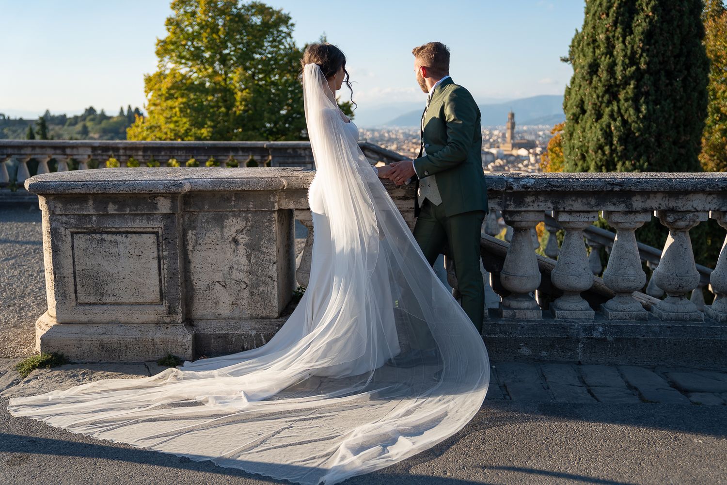 A couple in wedding attire shares an intimate moment on a stone balcony overlooking a scenic mountain landscape.