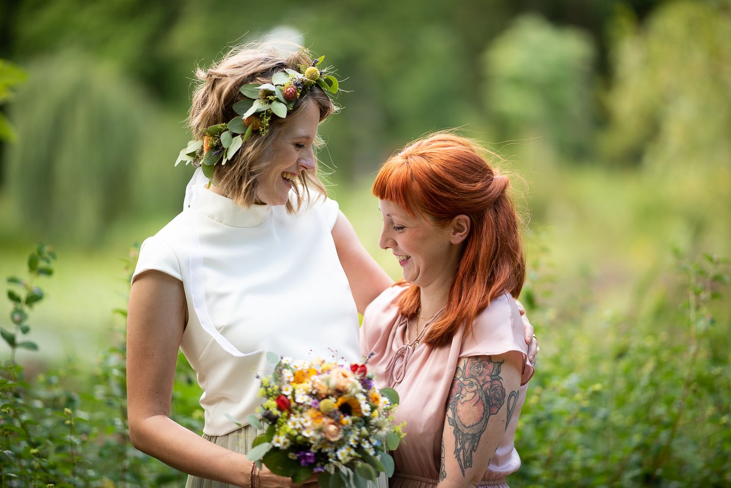 A couple embraces in a lush garden setting surrounded by greenery and holding bright orange and yellow flowers.