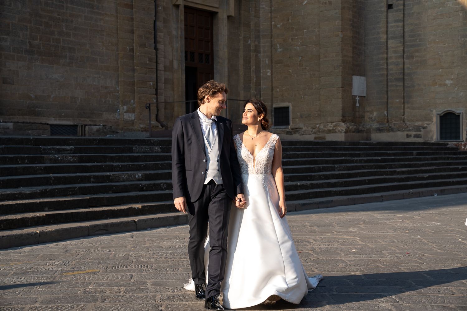A bride and groom walk along a grand stone building in sunlight, creating a romantic wedding scene.