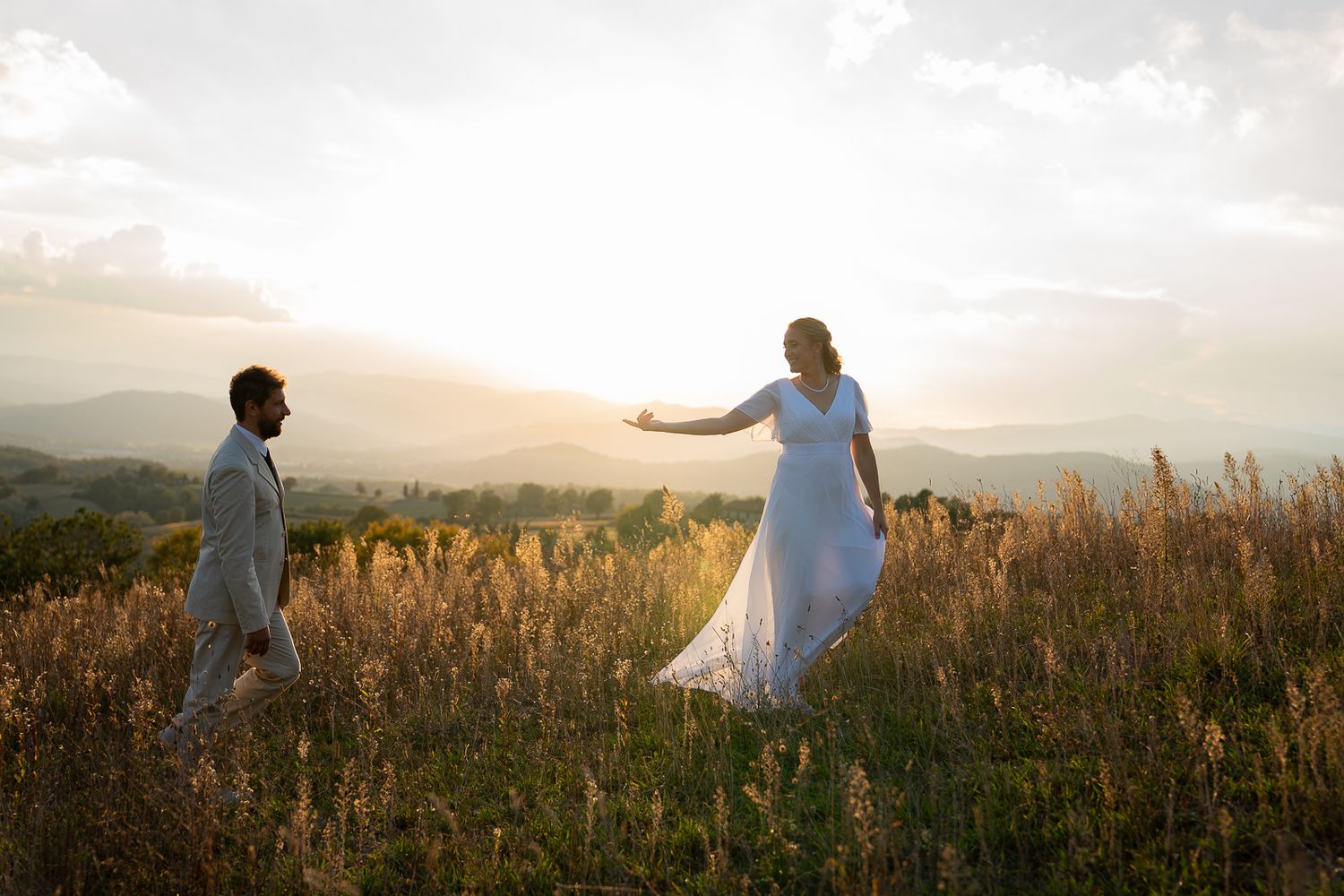 A couple in white attire dances through a golden field at sunset with mountains in the background near Montone, Umbria