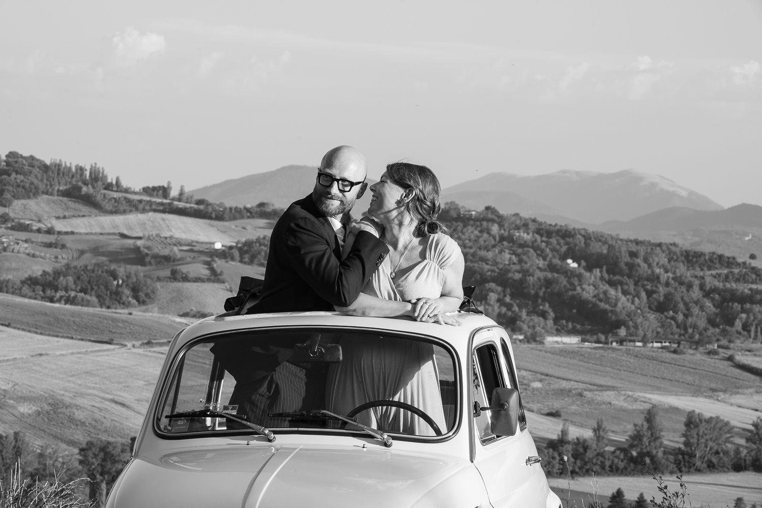 Black and white photograph of couple with vintage car in rural landscape.