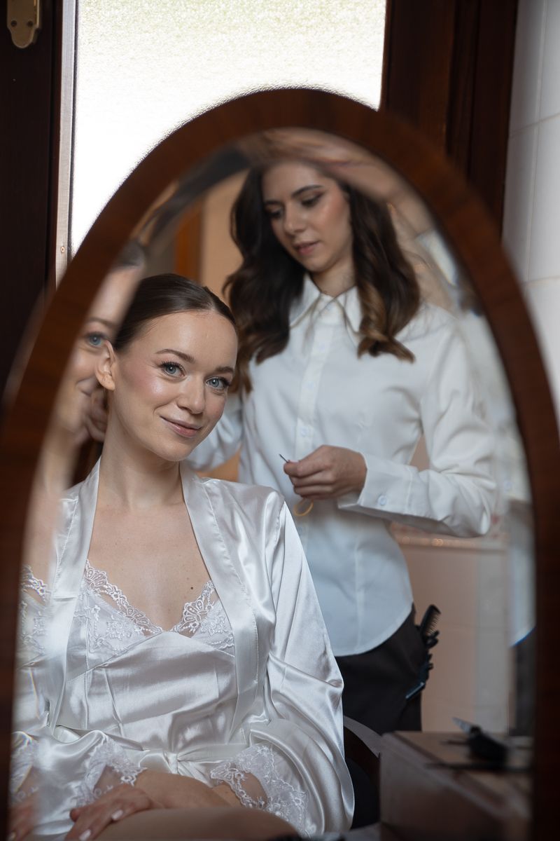 Bride in white satin robe sits before mirror as stylist applies makeup during wedding preparation.