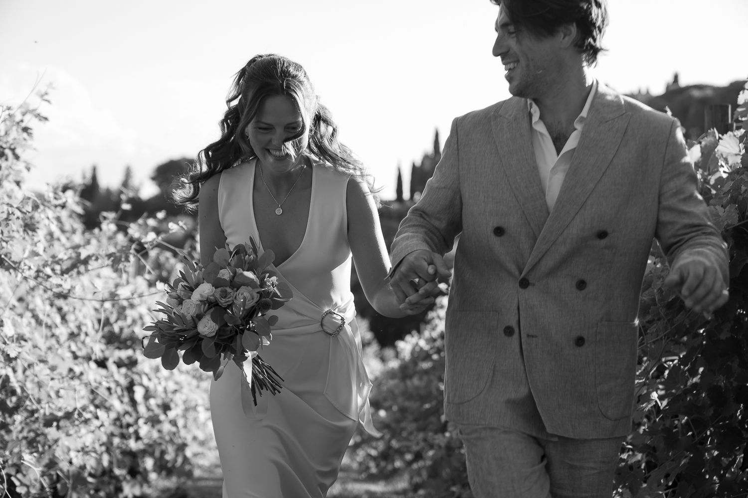 Black and white photo of a bride and groom holding hands walking through a vineyard, bride carrying a bouquet.