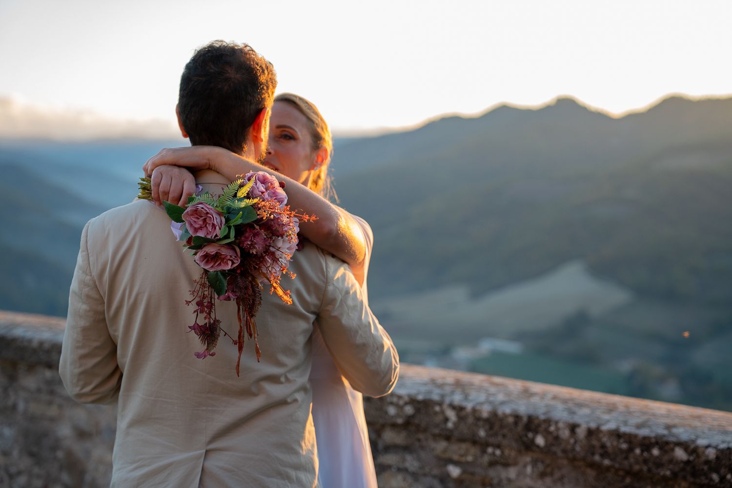 A couple embraces at sunset on a mountain overlook with a colorful bouquet in hand.