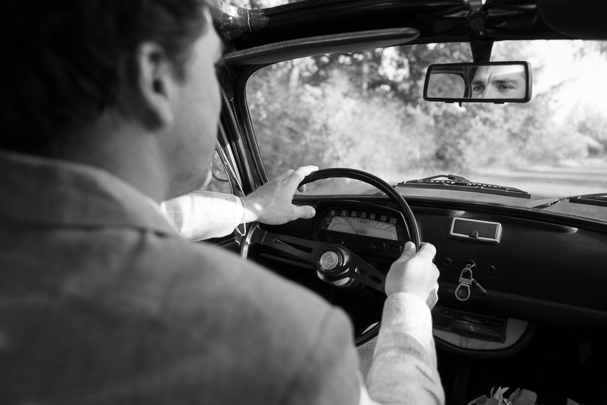 Black and white photo of a man driving a vintage car, hands on steering wheel, viewed from behind.