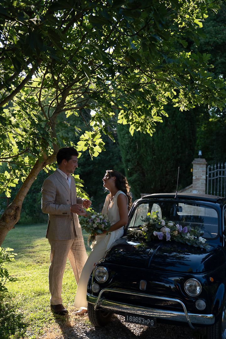 Bride and groom standing beside a vintage black Fiat 500 decorated with flowers in a sunlit garden.