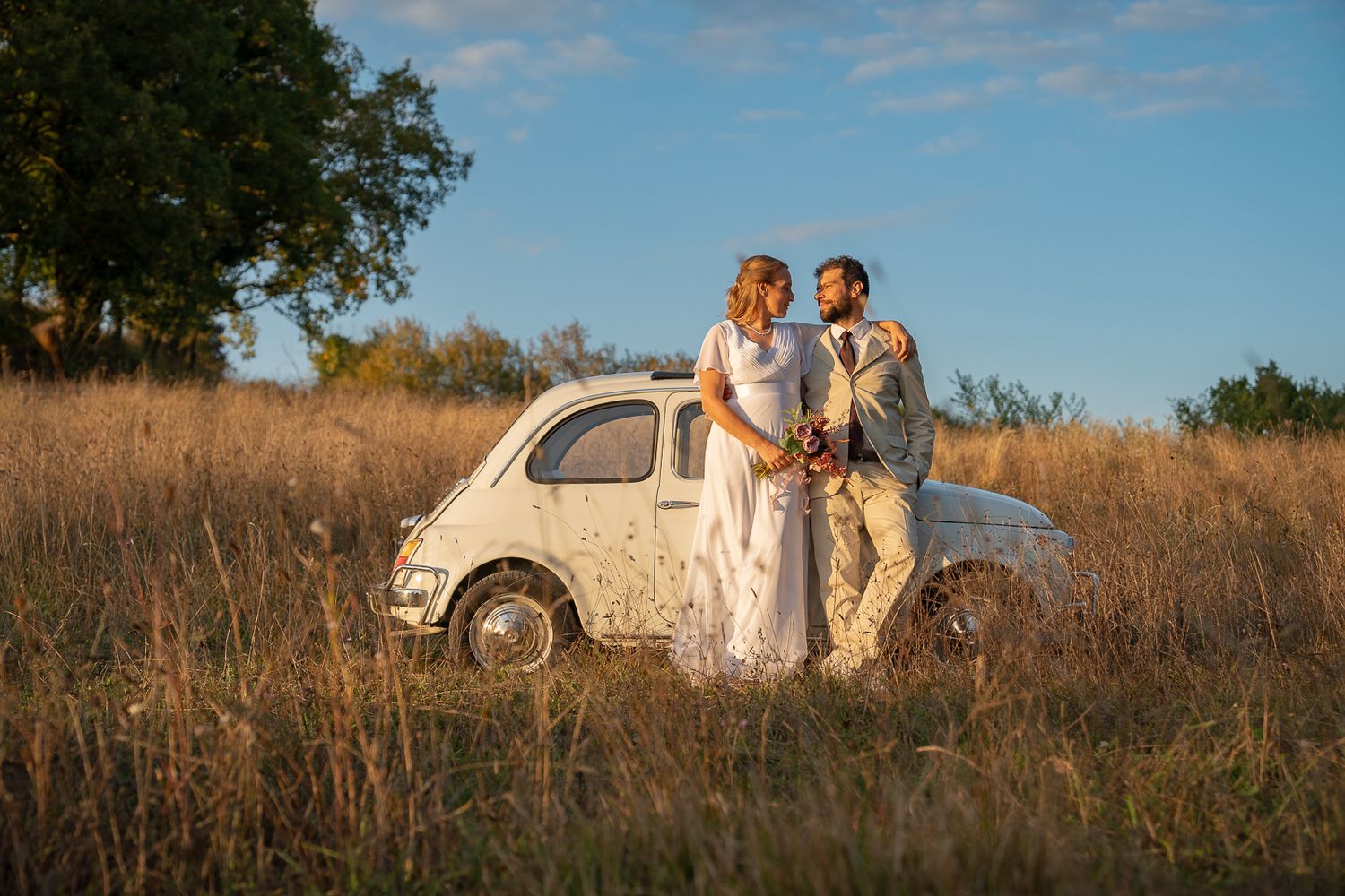 A vintage white Fiat 500 parked in a golden field at sunset with a couple in wedding attire standing beside it.