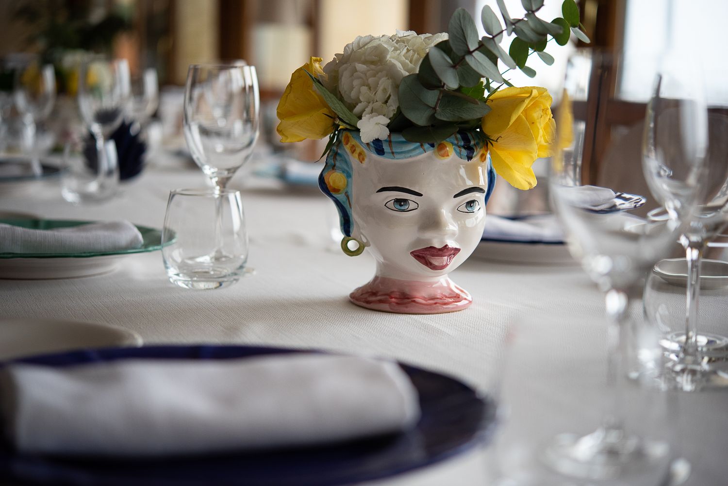 Decorative ceramic head vase with yellow flowers and foliage used as table centerpiece.