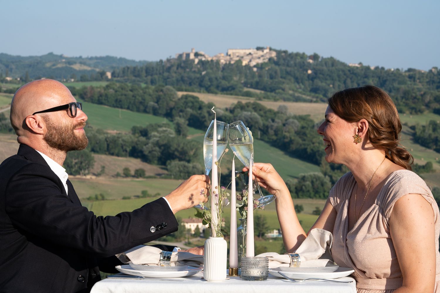 A couple toasts with champagne glasses at an elegant outdoor table setting overlooking the hills at Casale San Cristoforo, Italy