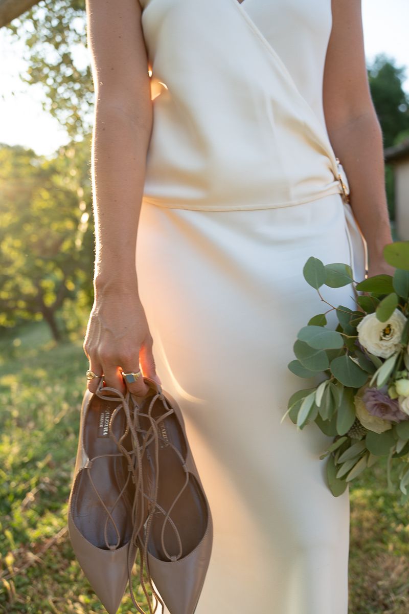Bride in ivory wedding dress holding nude lace-up flats and a green and white floral bouquet outdoors.