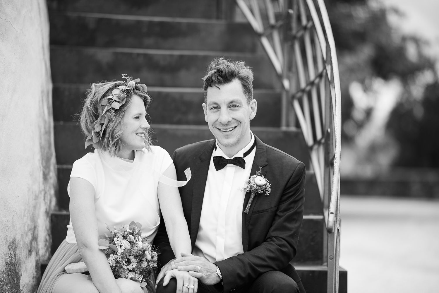 A couple shares a joyful moment sitting on outdoor steps in black and white wedding attire.