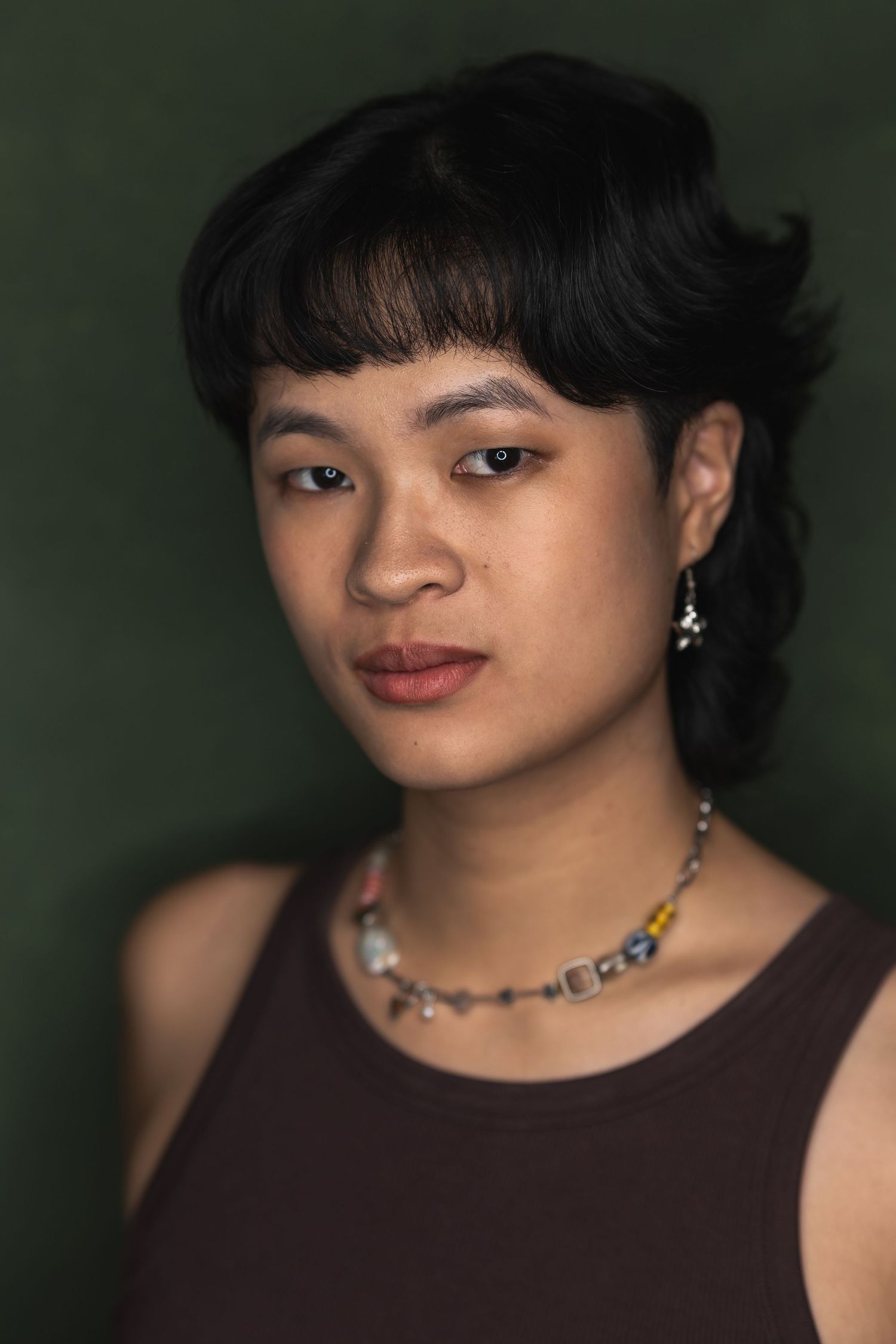 Portrait of someone wearing a dark sleeveless top and silver necklace against a dark green background.