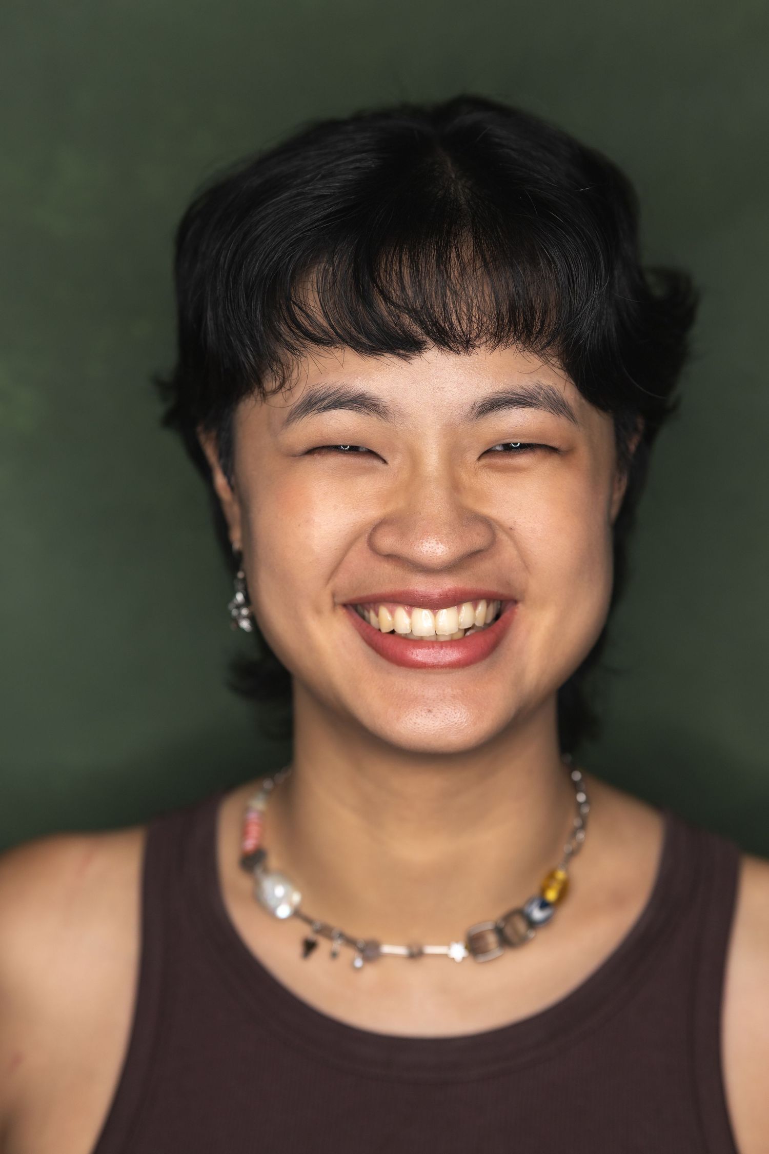 Close-up portrait showing a bright smile and silver jewelry against a dark green backdrop.