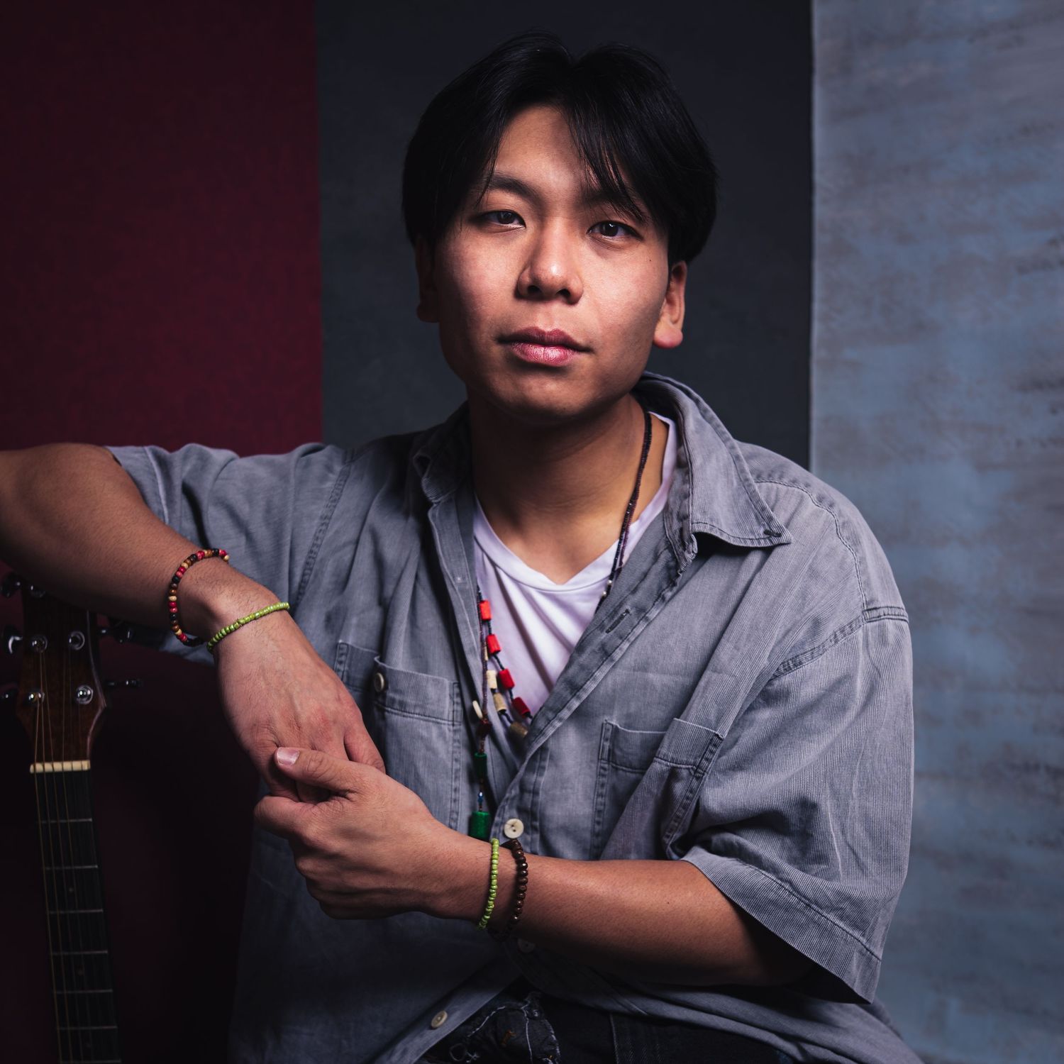 Portrait of a person in a gray button-up shirt leaning against a wall with a guitar visible in the background.