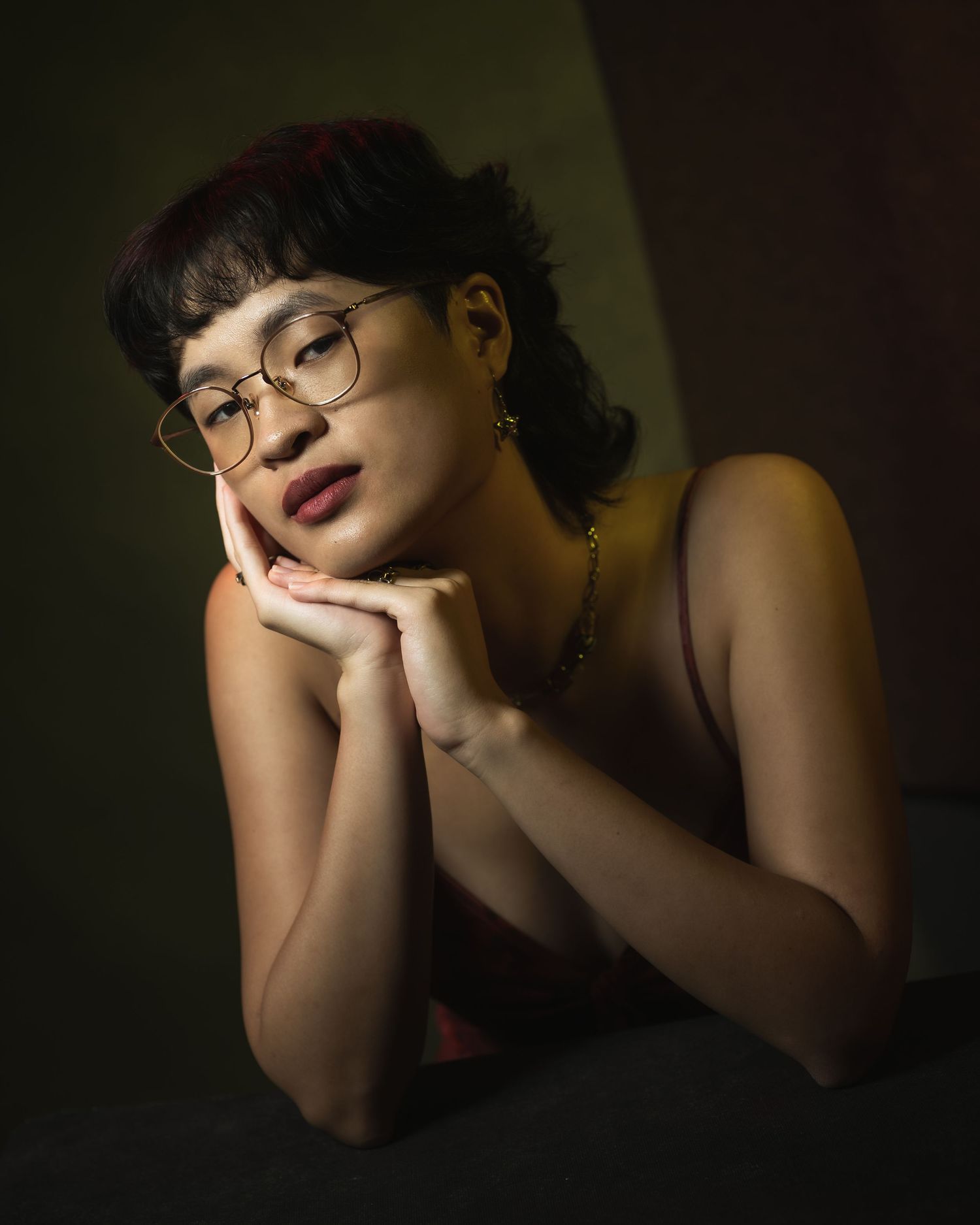 Portrait in moody lighting showing a contemplative pose with glasses and dark curly hair against a shadowy background.