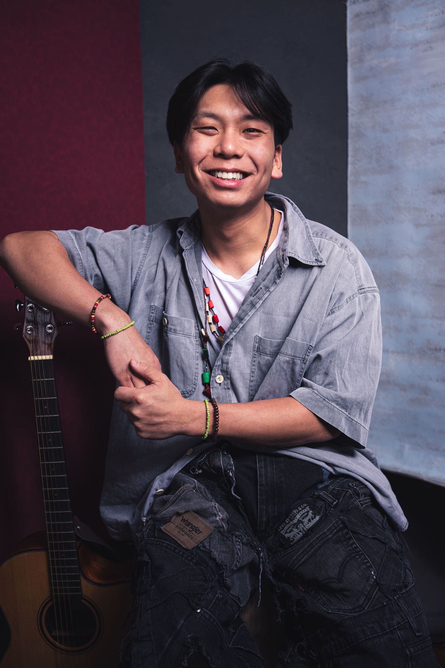 A young musician in a gray shirt poses with an acoustic guitar against a textured backdrop.