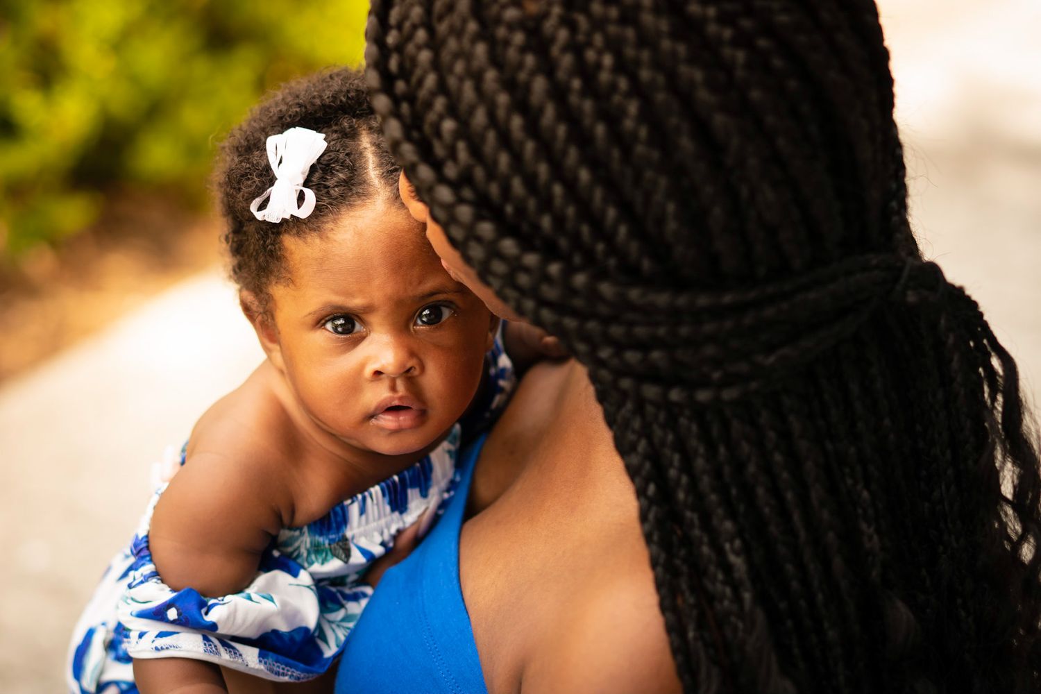 A small child wearing a blue and white dress sits with mother wearing braided hair.