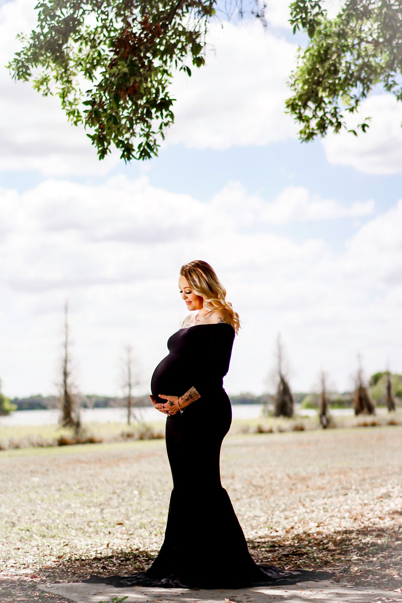 Silhouette of a person in black dress sitting outdoors under tree branches and cloudy sky.