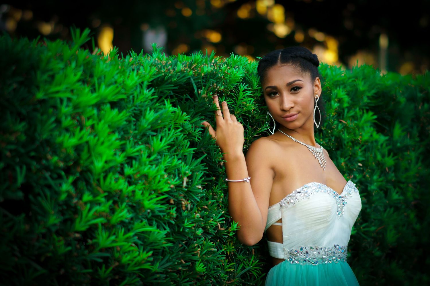 A person in a white formal dress poses elegantly in a lush garden setting with vibrant green foliage.