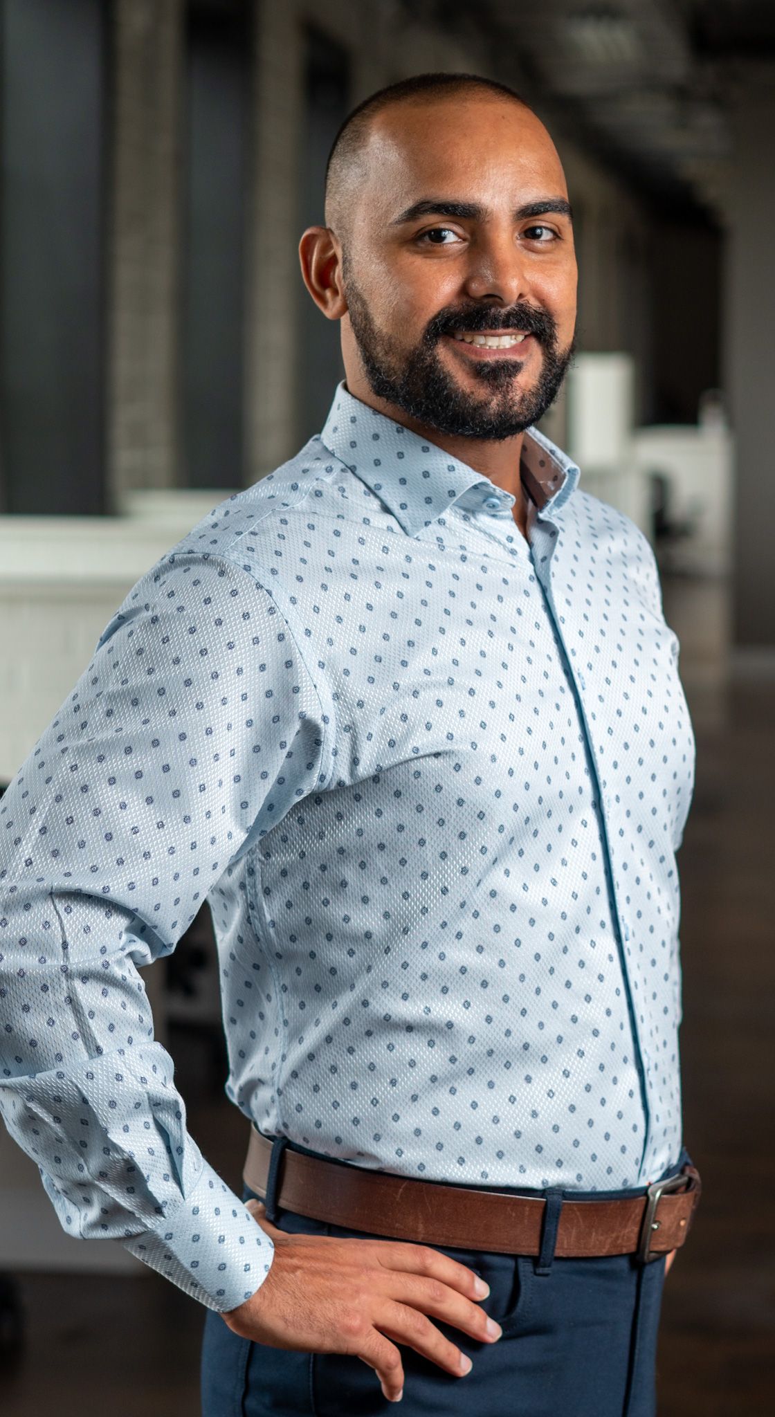 Man wearing white polka dot dress shirt with brown belt and blue pants poses in professional setting.