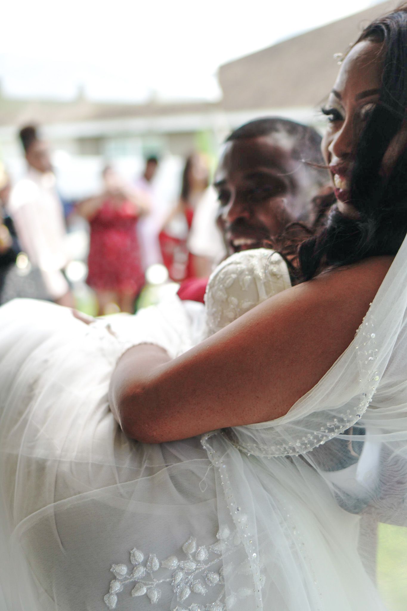 A bride in a white wedding dress being hugged at an outdoor celebration.