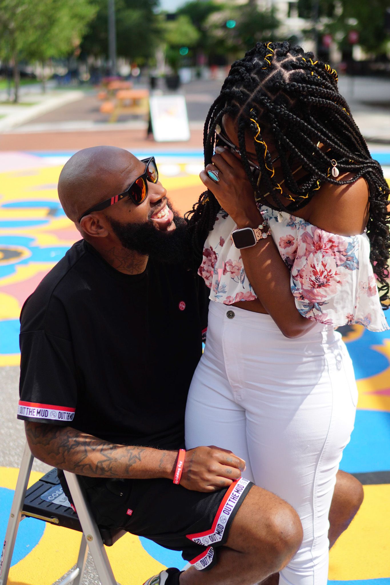 A couple in black and white outfits share an intimate moment outdoors with colorful artwork in the background.