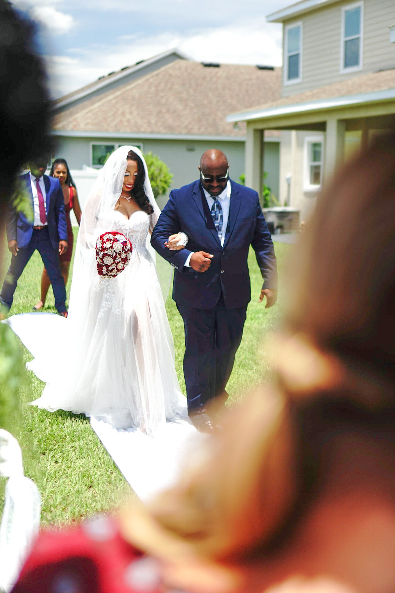 Wedding ceremony outdoors with white dress and navy suits against residential backdrop.