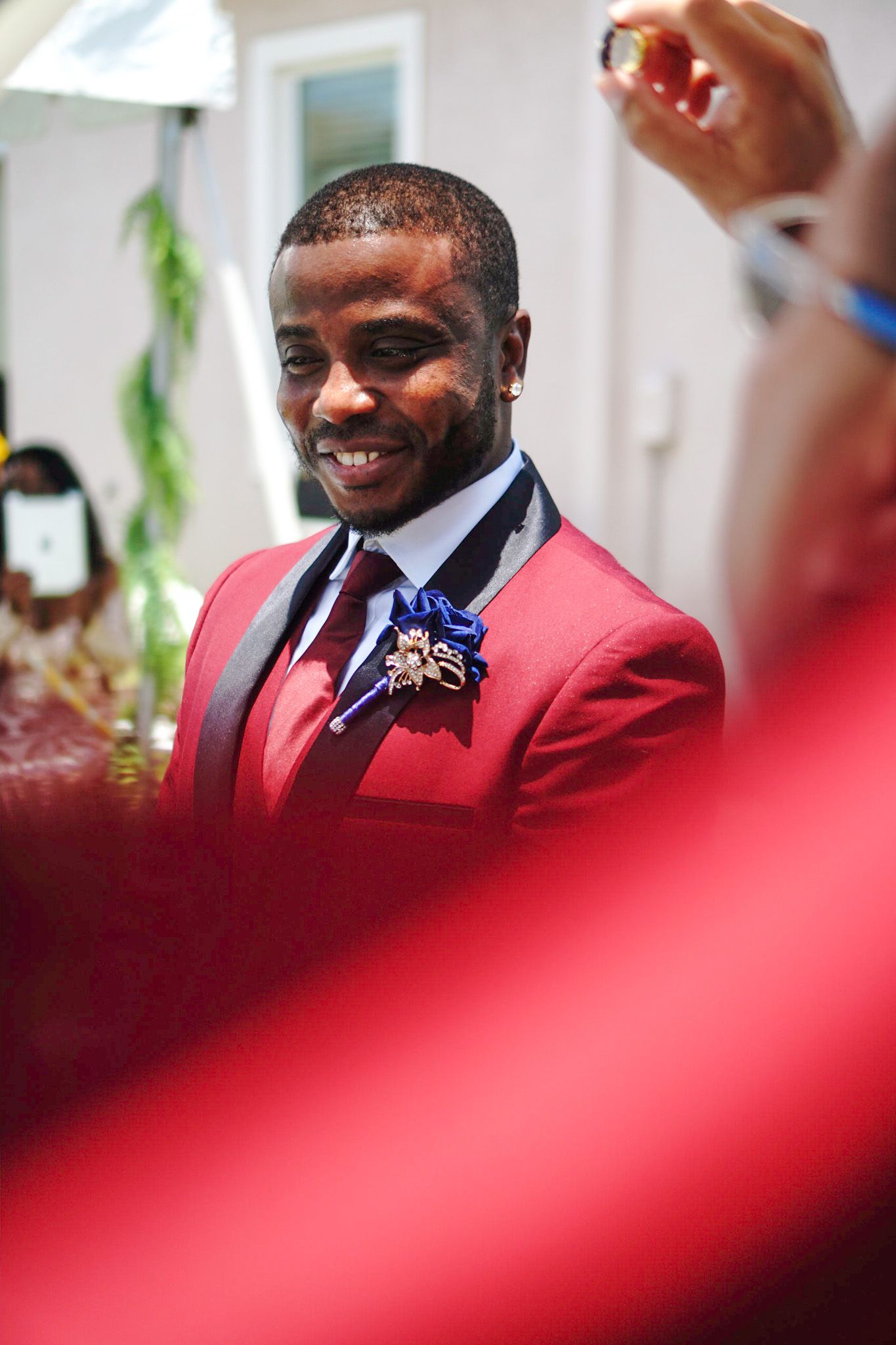 A person in a red suit jacket smiles warmly during a formal wedding event.