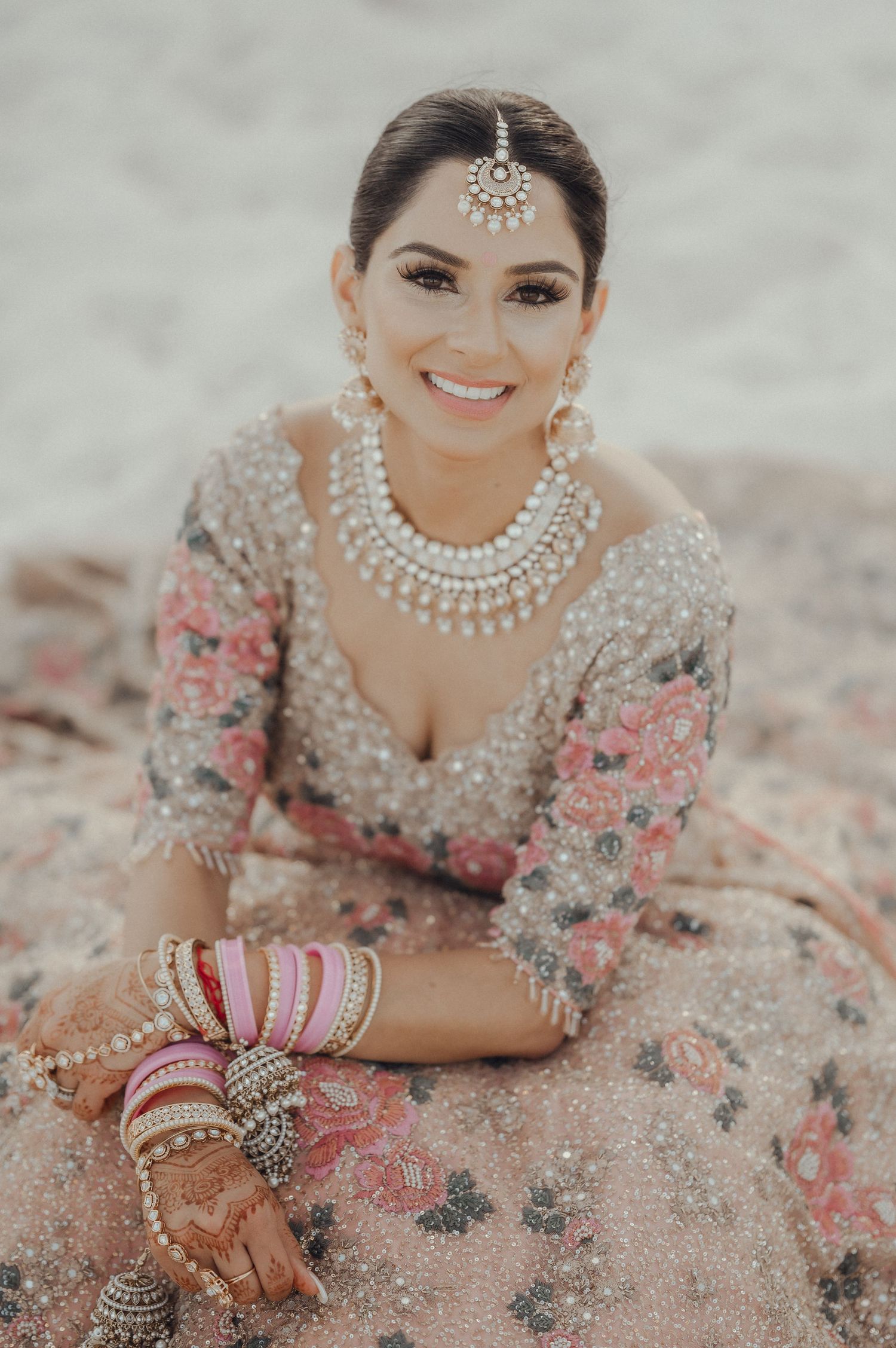Traditional Indian bridal attire with floral embroidered lehenga, statement jewelry and bangles in soft pink and silver tones.