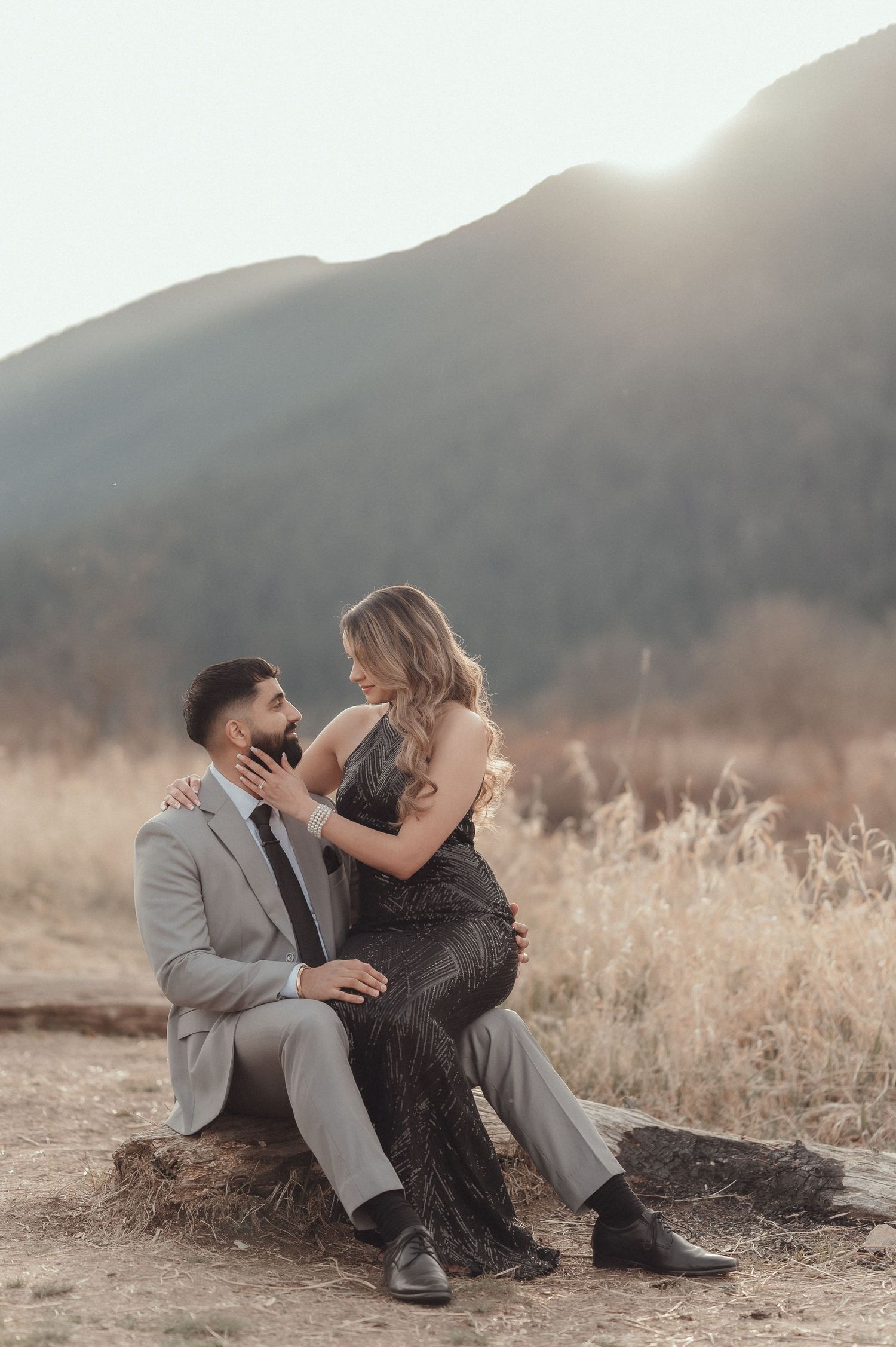 Couple in formal attire sharing playful moment against mountain backdrop at sunset.
