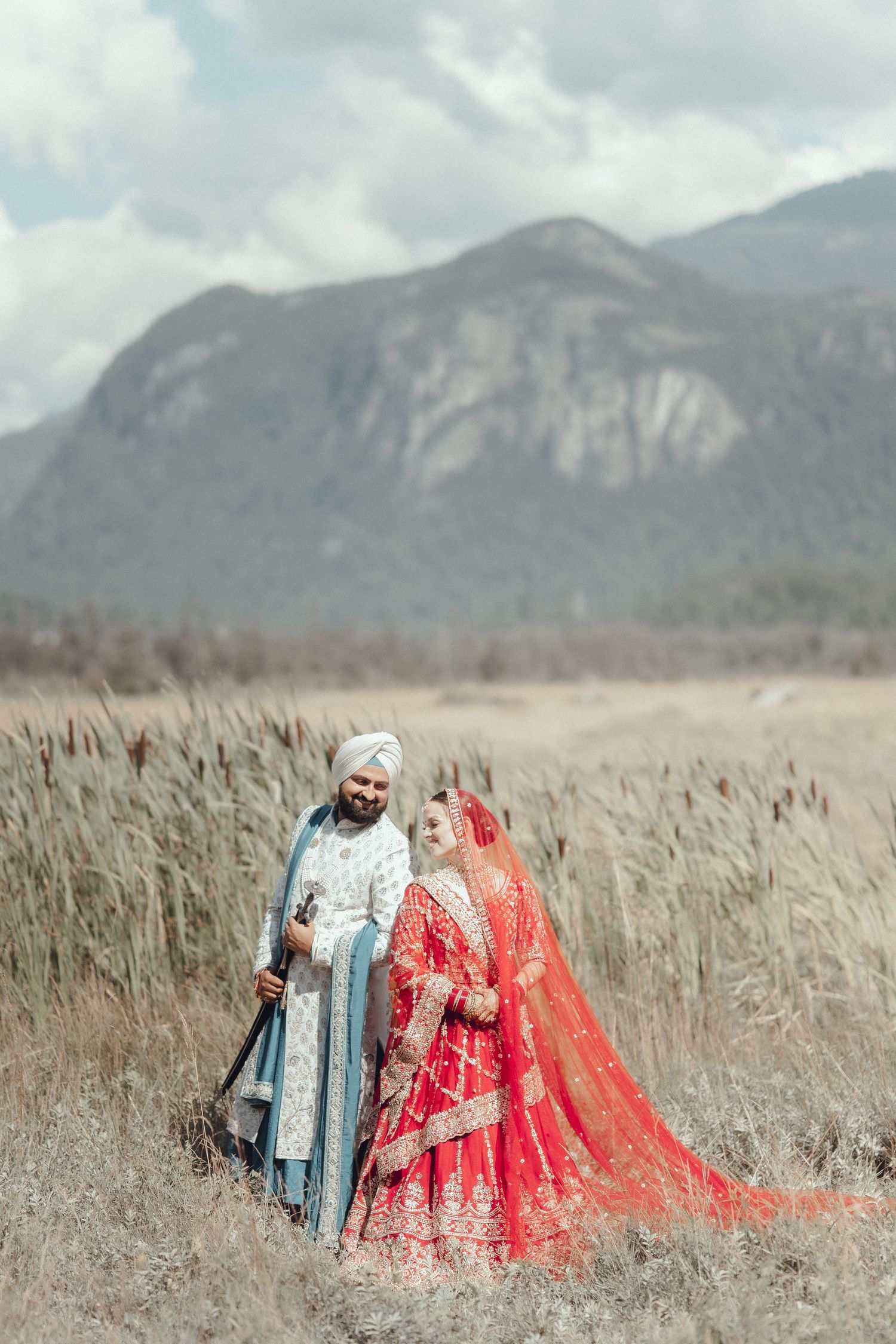 Romantic couple in traditional South Asian wedding attire pose together in a grassy mountain meadow with scenic peaks behind them.