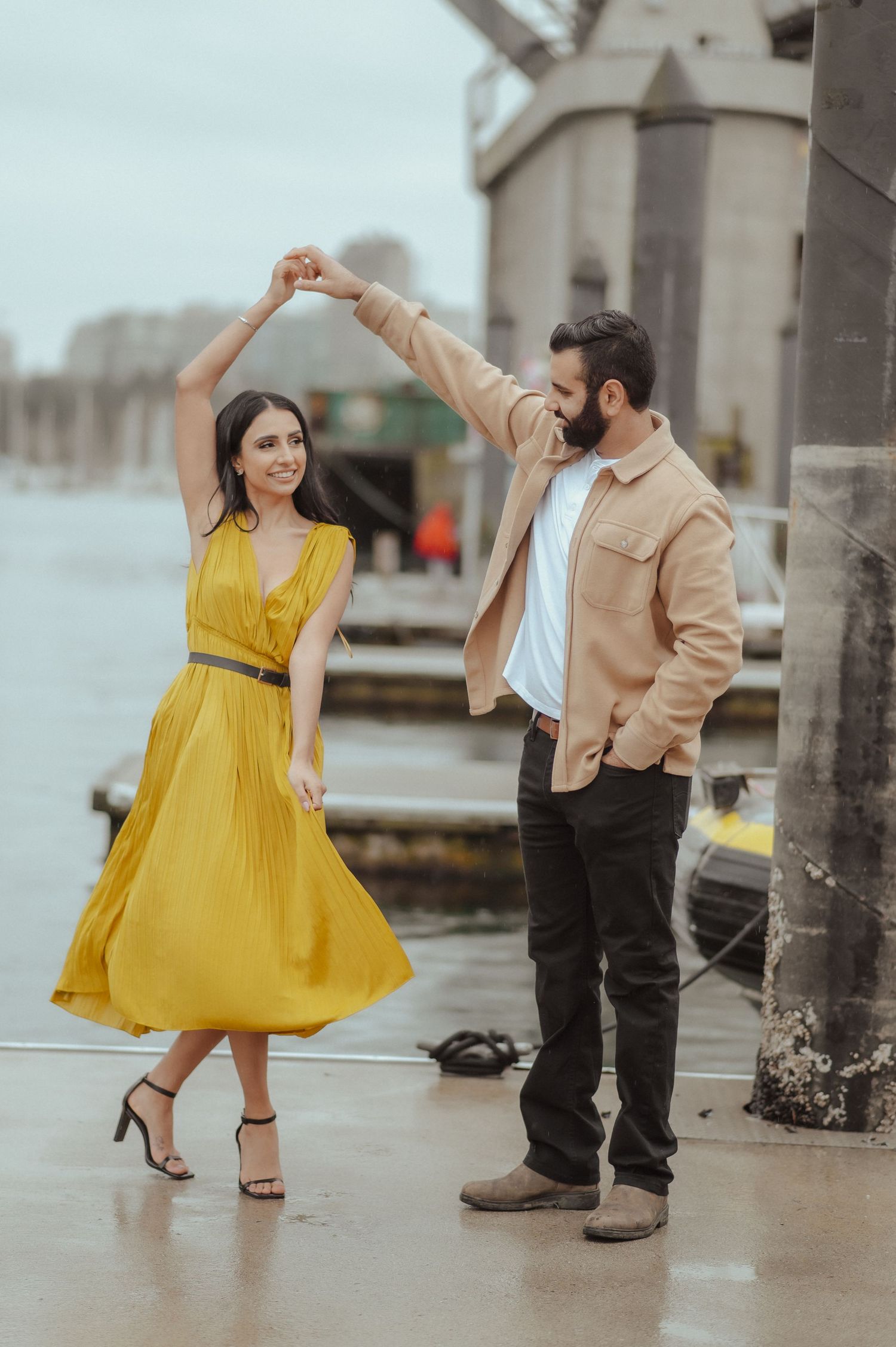 A couple dances together on a waterfront, with the woman wearing a bright yellow dress and the man in beige.