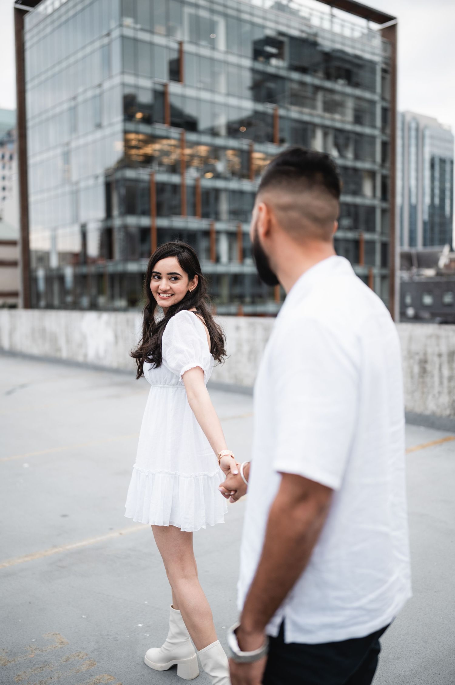 Couple in matching white outfits posing against modern glass building backdrop.