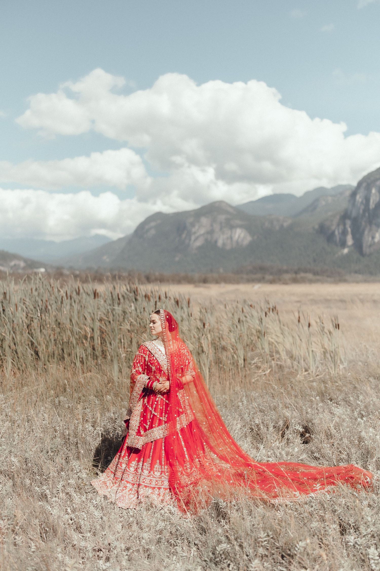 A flowing red wedding dress with long train photographed against a scenic mountain landscape in dried grass field.